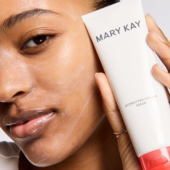 A hand holds a white tube of Mary Kay product with a pink cap, positioned against a softly lit background.
